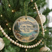 A round ornament hanging on a Christmas tree, featuring a scenic landscape with mountains, pine trees, and a lake.