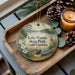 A decorative ornament with a scenic lake and park landscape, featuring a bird and a canoe, is displayed on a wooden tray alongside pine cones and a lit candle.