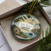 A decorative ornament featuring a scenic landscape with a lake, mountains, and a small boat, placed in a box with pine branches.
