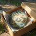 A decorative ornament in a box, featuring a scenic landscape with a lake, mountains, and a small cabin.