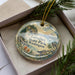 A decorative ornament in a box, featuring a scenic landscape with pine trees, mountains, and a bird perched on a branch.