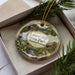 A decorative ornament in a box, featuring a scenic landscape with a house, trees, and a waterfall.