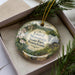 A decorative ornament in a box, featuring a scenic landscape with mountains, trees, and a waterfall.