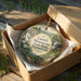 A decorative ornament in a box, featuring a scenic landscape with mountains, trees, and a waterfall.