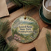 A circular ornament with a scenic mountain landscape and a bird is displayed on a wooden surface, surrounded by pine branches and a cup of coffee.