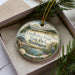 A decorative ornament in a box, featuring a scenic landscape with pine trees, a body of water, and a bird.