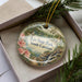 A decorative ornament in a box, featuring a scenic landscape with a waterfall and a bridge.