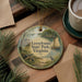 A round ornament with a painting of a sailboat and a bird, surrounded by pine branches and a cup of coffee on a wooden surface.