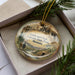 A decorative ornament in a box, featuring a scenic landscape with mountains, pine trees, and a person walking along a trail.
