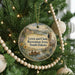 A round ornament hanging on a Christmas tree, featuring a scenic landscape with a river, mountains, and people in traditional clothing.
