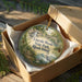 A decorative ornament in a box, featuring a scenic landscape with a waterfall and greenery, with the text "Lincoln Woods State Park, Rhode Island" printed on it.