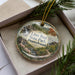 A decorative ornament in a box, featuring a scenic landscape with a waterfall and pine trees.