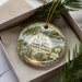 A decorative ornament with a nature scene, featuring a bird, flowers, and a path, is displayed in a box with pine branches.