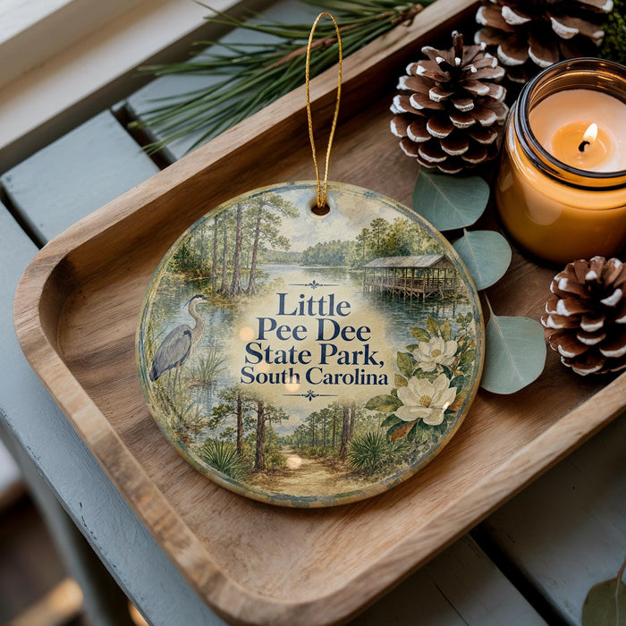 A decorative ornament featuring a scenic image of Little Pee Dee State Park, South Carolina, with pine cones and a lit candle on a wooden tray.