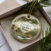 A decorative ornament in a box, featuring a scenic landscape with a house, trees, and a bridge.