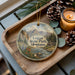A wooden tray with a decorative ornament featuring a scenic landscape of mountains, pine trees, and a river, along with pine cones and a lit candle.