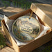 A circular ornament in a wooden box, featuring a scenic landscape with mountains, a river, and wildflowers.