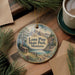 A decorative ornament featuring a scenic landscape with mountains, pine trees, and a cabin, hanging on a wooden surface.