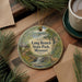 A decorative ornament featuring a scenic landscape with a waterfall, surrounded by pine branches and a deer, placed on a wooden surface next to a cup of coffee.