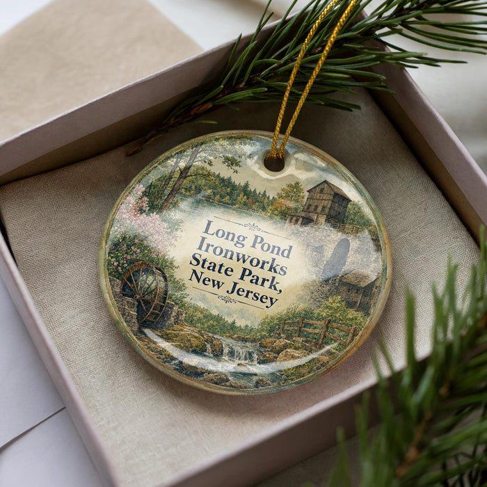 A decorative ornament in a box, featuring a scenic landscape with a pond, ironworks, and a building, surrounded by pine branches.