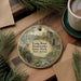 A round ornament with a painting of a scenic landscape, featuring a pond, ironworks, and a building, is displayed on a wooden surface alongside a cup of coffee and some pine branches.