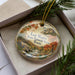A decorative ornament in a box, featuring a scenic landscape with mountains, trees, and a river.