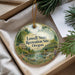 A round ornament with a scenic landscape featuring a river, bridge, and trees, hanging from a string.