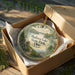 A decorative ornament in a wooden box, featuring a scenic landscape with a lake and trees, with the text "Madison Lake State Park, Ohio" printed on it.