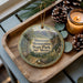 A wooden tray with a decorative ornament featuring a scenic mountain landscape and pine cones, along with a lit candle and pine cones.