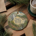 A round ornament featuring a scenic landscape with a waterfall, surrounded by pine branches and a cup of coffee on a wooden surface.