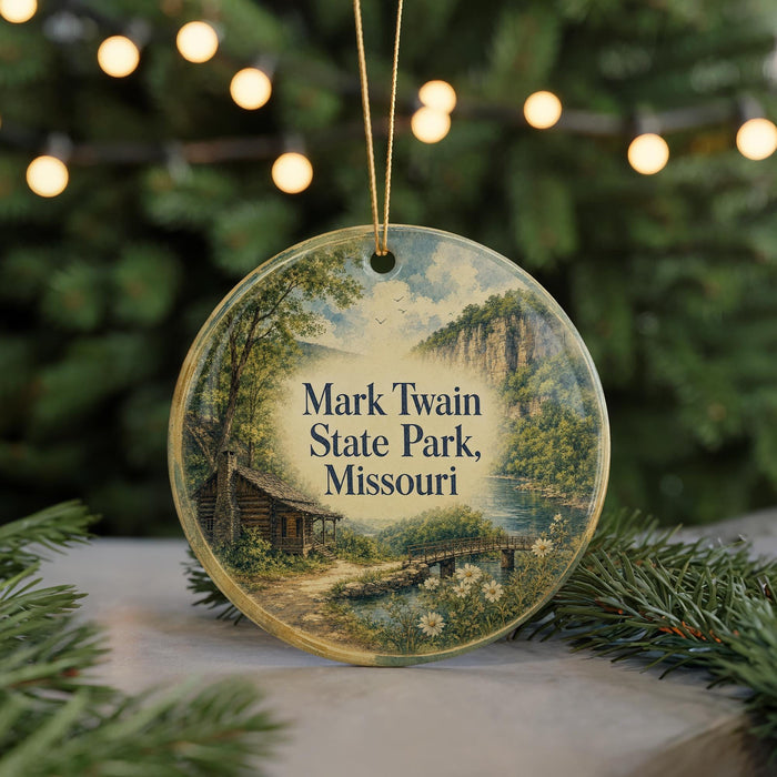 A round ornament with a painting of a cabin and a river, surrounded by greenery, hangs in front of a blurred background with twinkling lights.