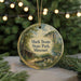 A round ornament with a painting of a cabin and a river, surrounded by greenery, hangs in front of a blurred background with twinkling lights.