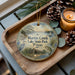 A decorative ornament with a painting of Martin Creek Lake State Park, Texas, sits on a wooden tray alongside pine cones and a lit candle.