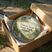 A decorative ornament in a brown box, featuring a scenic landscape with a lake, trees, and wildlife, including a bird and a deer.