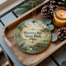 A decorative ornament with a serene scene of a lake, a lighthouse, and birds, along with the text "Maunee Bay State Park, Ohio". The ornament is placed on a wooden tray, with pine cones and a lit candle nearby.
