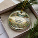 A decorative ornament in a box, featuring a scenic landscape with a deer, trees, and a waterfall.