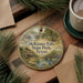 A decorative ornament featuring a scenic image of McKinney Falls State Park, Texas, with a small house and a waterfall in the background. The ornament is placed on a wooden surface, surrounded by pine branches and a cup of coffee.