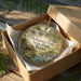 A circular ornament in a wooden box, featuring a scenic landscape with a waterfall and a house, with the text "McKinney Falls State Park, Texas" printed on it.