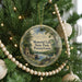 A round ornament hanging on a Christmas tree, featuring a scenic landscape with a lake and a stone bridge.