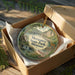 A decorative ornament in a box, featuring a scenic landscape with a river, bridge, and trees.