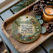 A decorative ornament with a scenic image of Mill Pond State Park in North Carolina, featuring a bird, a boat, and a cabin, is displayed on a wooden tray alongside pine cones and a lit candle.