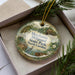 A decorative ornament in a box, featuring illustrations of a mill pond, a wooden cabin, and a bird, with the text "Merchants Millpond State Park North Carolina" printed on it.