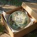 A decorative ornament in a wooden box, featuring a scenic landscape with a waterfall and forest.