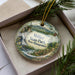 A decorative ornament in a box, featuring a scenic landscape with a waterfall, pine trees, and mountains.