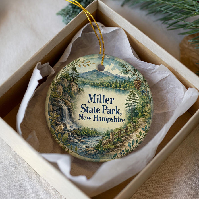 A decorative ornament in a box, featuring a scenic landscape with a waterfall and pine trees, with the text "Miller State Park, New Hampshire" displayed on it.