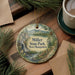 A decorative ornament featuring a scenic landscape of Miller State Park in New Hampshire, with pine trees and a waterfall, is displayed on a wooden surface alongside a cup of coffee and some papers.