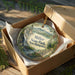 A decorative ornament in a brown box, featuring a scenic landscape with a waterfall and pine trees, with the text "Miller State Park, New Hampshire" printed on it.