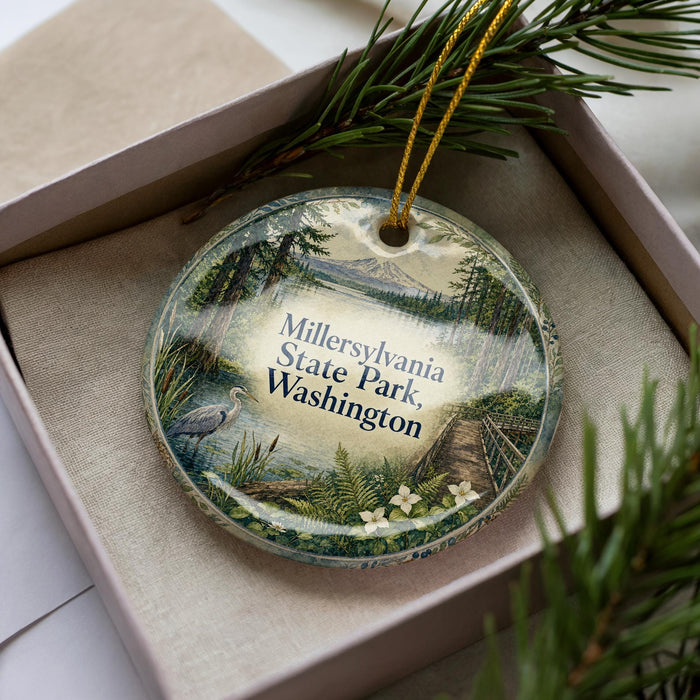 A decorative ornament in a box, featuring a scenic landscape with a lake, mountains, and pine trees.