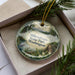 A decorative ornament in a box, featuring a scenic landscape with a lake, mountains, and pine trees.