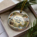 A clear ornament with a landscape scene featuring a river, mountains, and a building, hanging from a string.